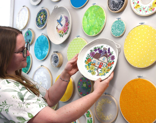 Woman holding an embroidered rope basket with house and florals on it. Surrounded by other embroidered rope baskets and hoops.